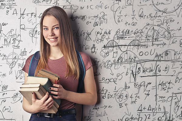 Girl holding a stack of books and standing in front of a wall filled with equations