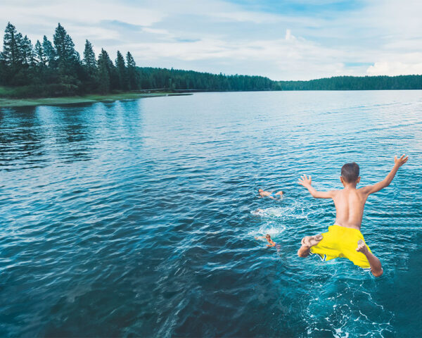 The expanded image show the same leaping boy, now wearing yellow shorts. The lake contains only three other boys, but we can see the distant shorline with pine trees and partly cloudy blue sky above.