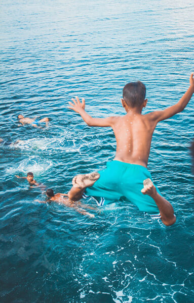 Source image showing a boy in squa blue shorts leaping into the water. We're looking from behind an over the boy, and see only water and four other swimmers in the frame.